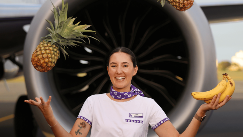 Bonza flight attendant juggling fruit in front of Bonza aircraft.